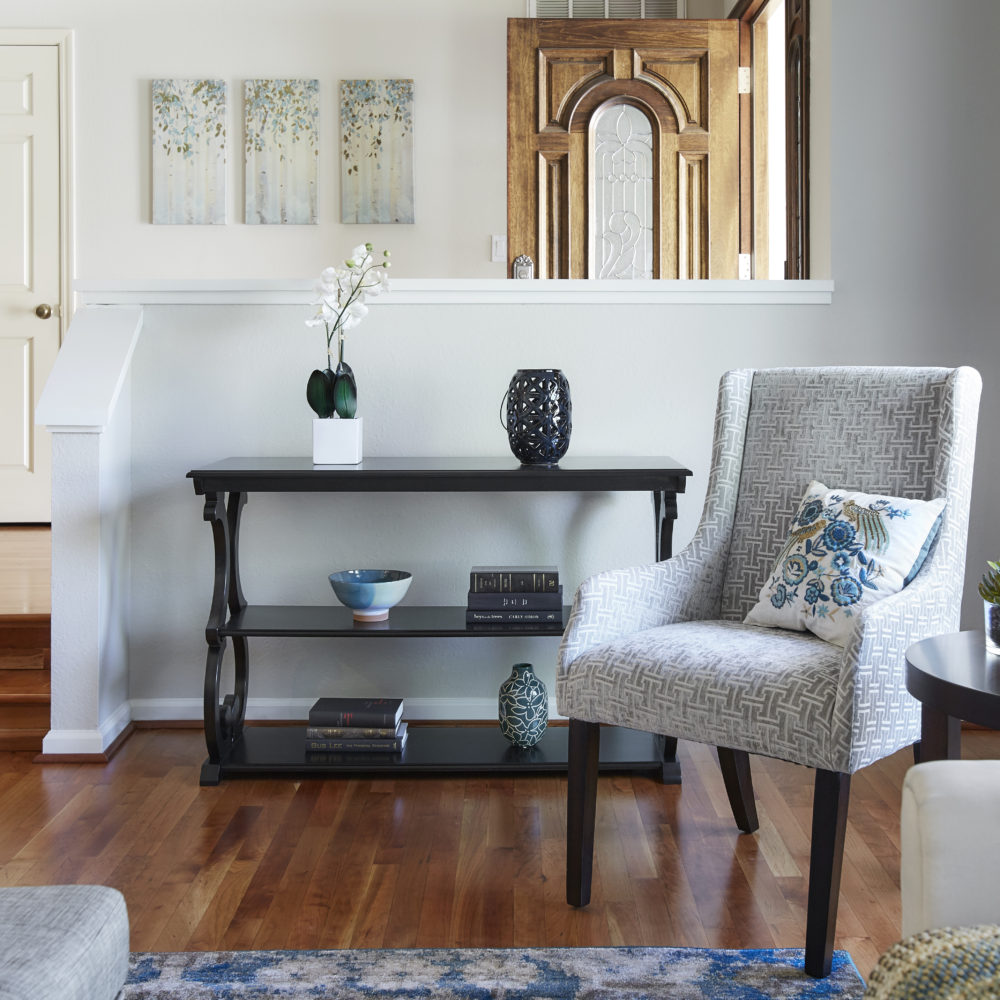 In another angle of our formal living room, we see our antique black scroll sofa table, decorated with various pieces of décor. Next to the table is one of the grey and white linen upholstered sloped arm accent chairs.