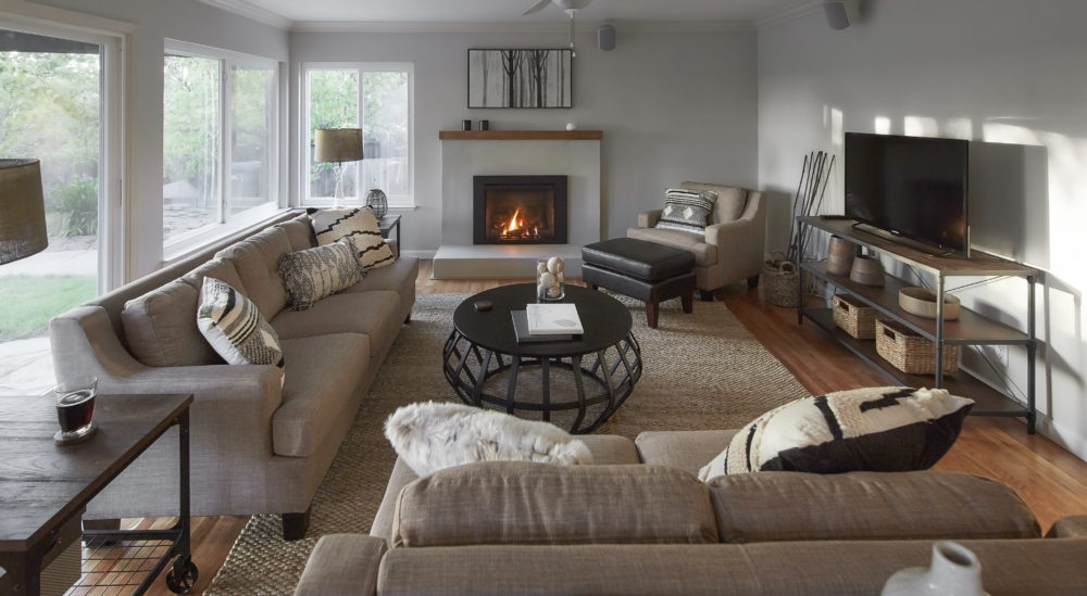 This is the "after" image of the casual living room. The white tiled floors are replaced with hardwood, the walls are pained white, and the fireplace has been completely remodeled. The room features a grey fabric sofa, grey fabric loveseat, black cage drum coffee table, grey fabric accent chair, brown faux leather footstool, and a wood and metal TV stand with a TV on it.