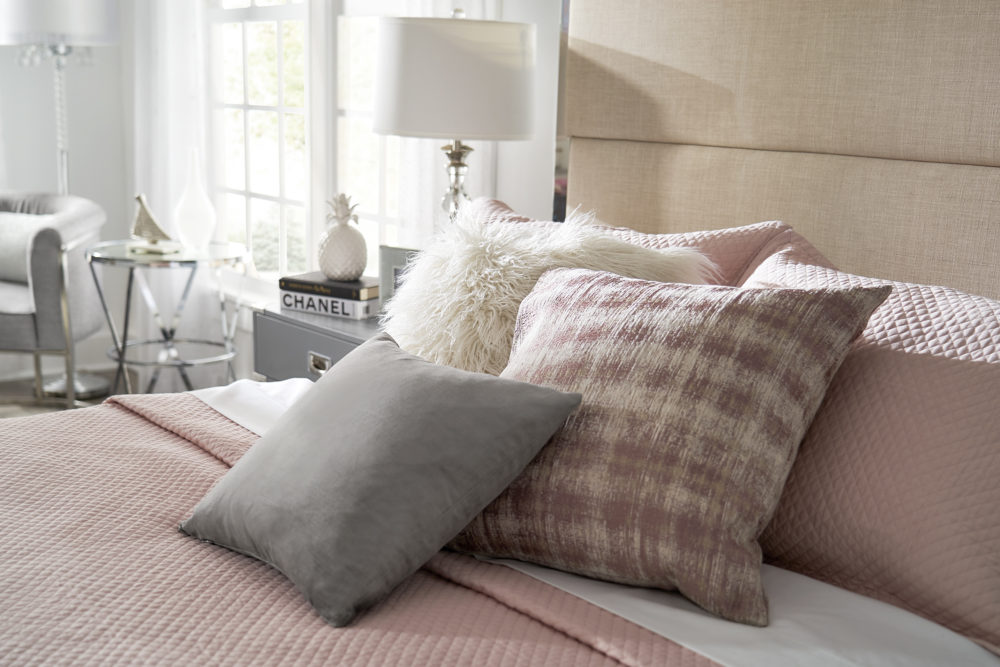 This photo is a closeup of the bedding, so you can see the pink blanket and pillows of pinks, whites, and greys. The background you can see the grey campaign nightstand as well as a round chrome end table.