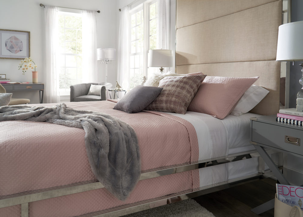This is another angle of our canopy bed. It shows the pink, grey, and white bedding that pairs beautifully with the chrome metal frame and beige upholstered headboard. It also is complimented by the grey campaign nightstands, grey campaign writing desk, and grey linen upholstered accent chair.