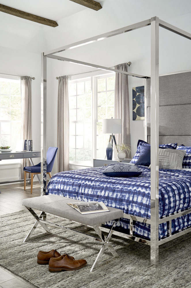 This is the second of our canopy bed ideas: masculine. This shows the same canopy bed with chrome metal frame, but it has a grey linen upholstered headboard. The bedding is blue, black, and grey. At the foot of the bed is a grey linen upholstered bench. There is also a grey campaign nightstand, grey campaign writing desk, and twilight blue barrel back linen upholstered side chair.