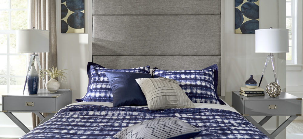This is a front view of the masculine-styled canopy bed and the two nightstands. The bed features blue, white, grey, and black bedding. The campaign nightstands are decorated with table lamps and modern pieces of décor.