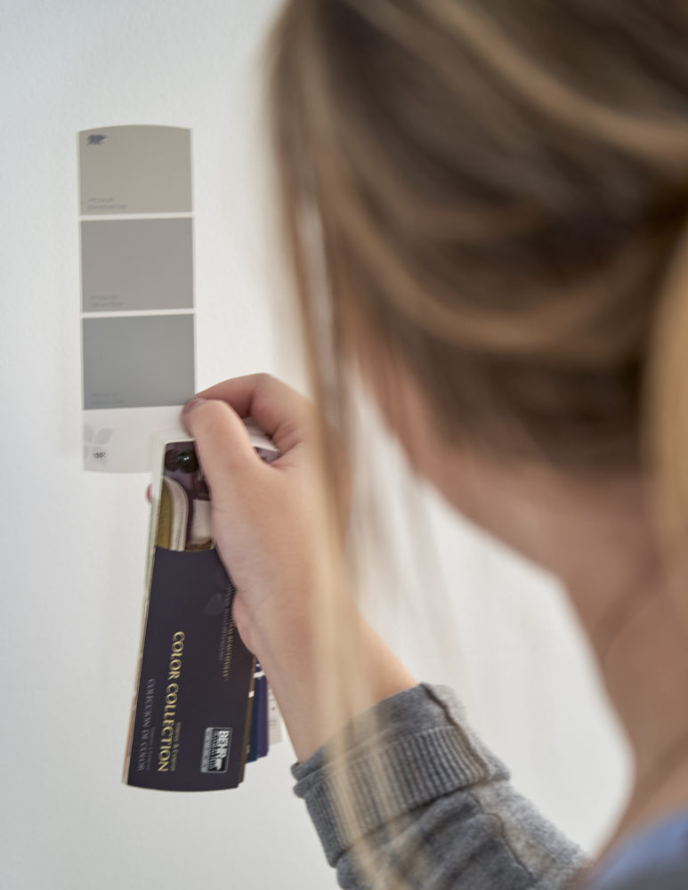 Before we begin the last of our canopy bed ideas, we decided to repaint our walls! This image shows our designer holding up a paint swatch of shades of grey to a white wall.