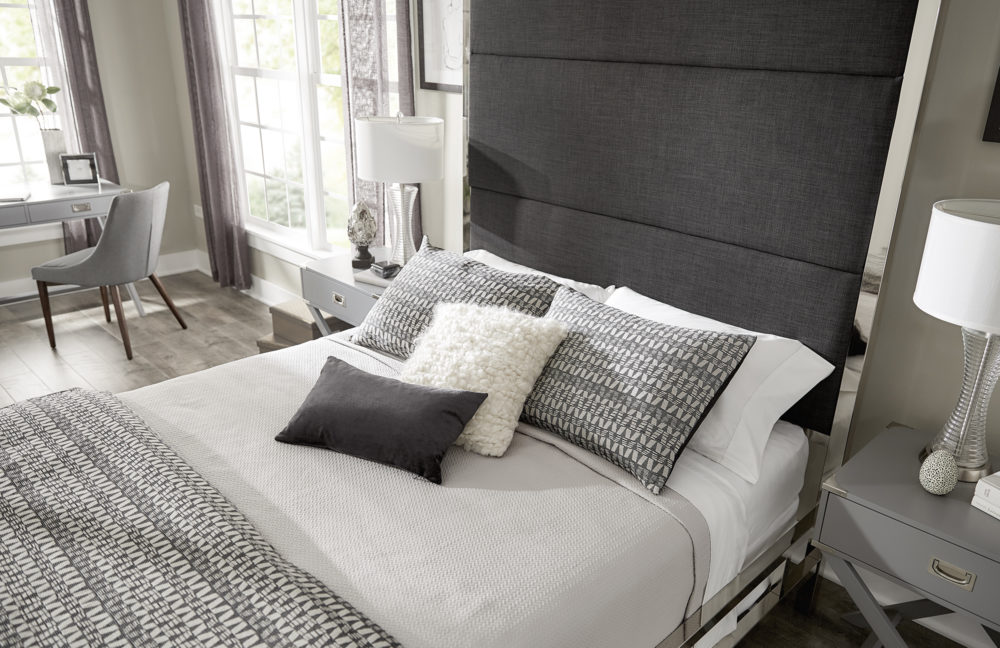 This image shows the neutral bedding from a higher angle so you can see the different patterns and textures of the black, white, and grey pillows and blankets. The grey campaign nightstands each have a table lamp and modern-themed décor. In the background, we have the grey finish campaign writing desk with a grey linen upholstered barrel back side chair.
