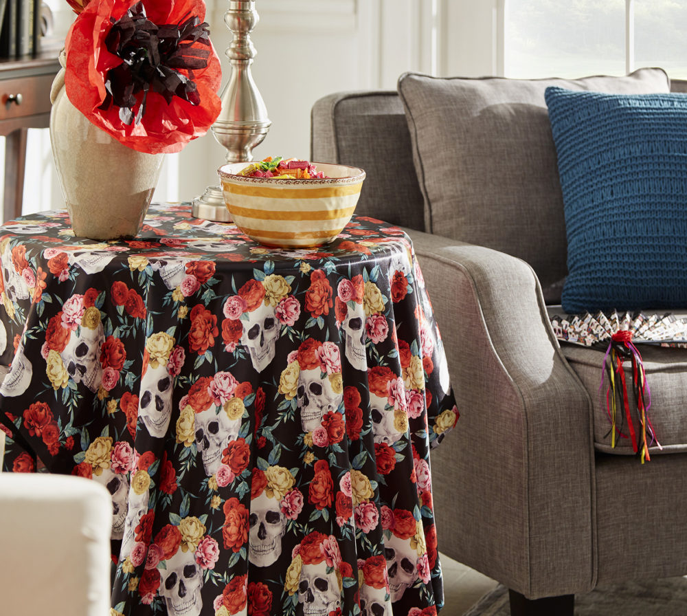 Another one of our Day of the Dead décor ideas is decorating an end table with a Day of the Dead-themed table cloth, a bowl of candy, and a vase with a paper poppy.