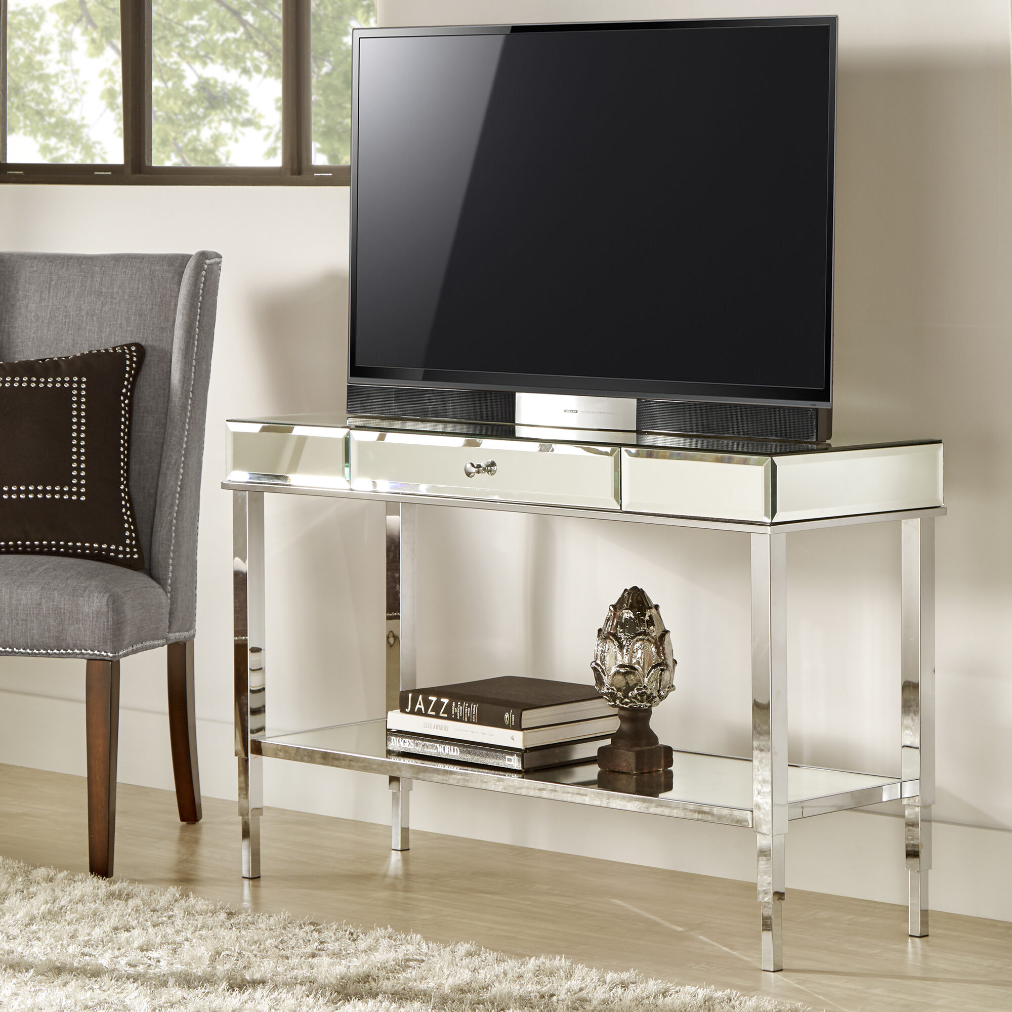 No matter which of our family room ideas you go with, always be sure to show off your style! This is our Camille Mirrored TV Stand Console Table with Drawer by iNSPIRE Q Bold. A flat screen TV sits atop a mirrored surface, while books and a piece of home decor sits on the lower open mirrored shelf. 