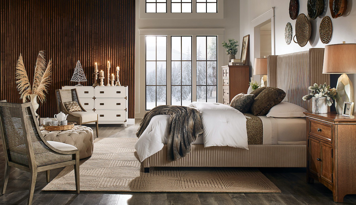 Pictured is our bedroom with all our home decor trends for 2021. The center of the room has a taupe velvet upholstered bed with two wood nightstands. The room also features a tall wood chest, a white and gold dresser, a beige button tufted cocktail ottoman, and two antique grey wood accent chairs.