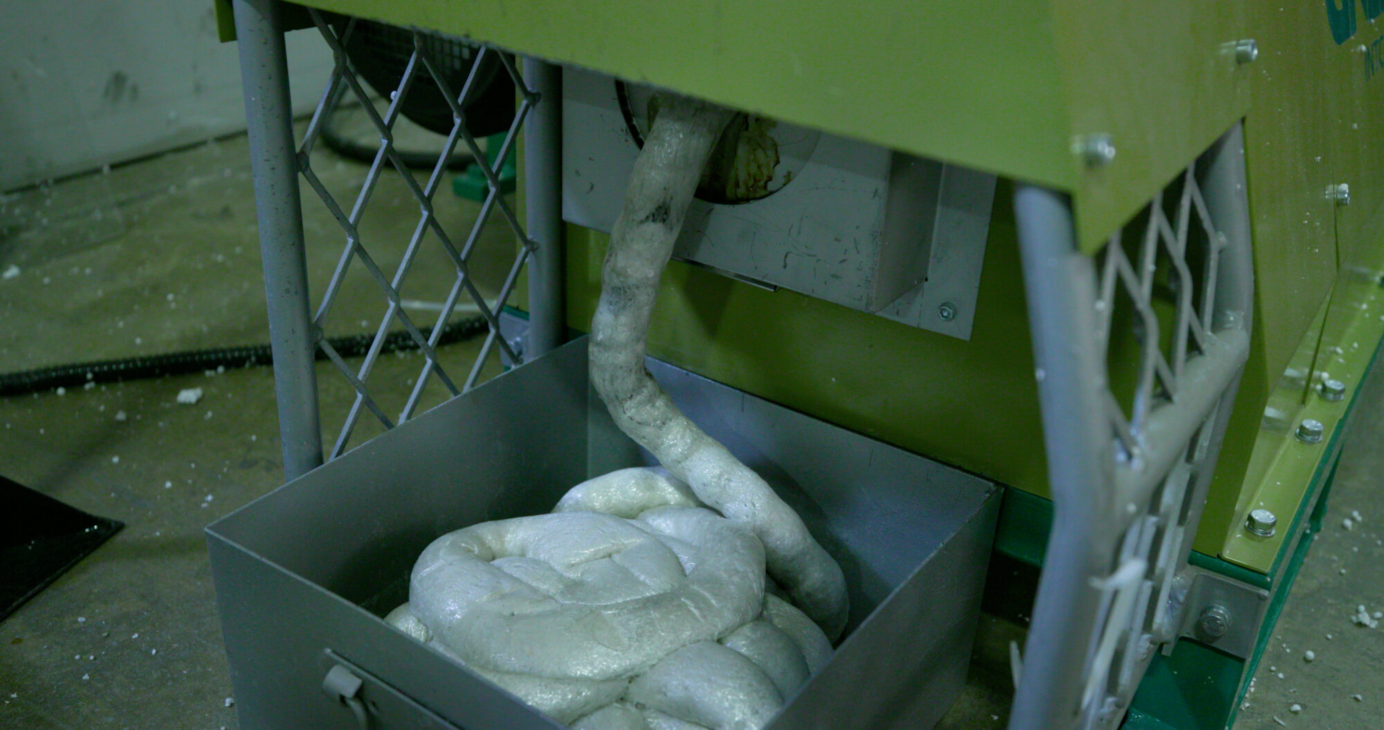 A closeup of the side of the GREENMAX machine. The melted white Styrofoam flows out of the machine into a metal box. 