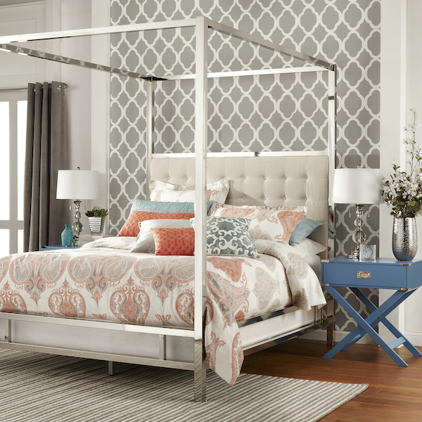 This modern canopy bed has a beige linen-upholstered headboard with plush button tufting. The metal frame is finished in chrome, which complements the beige linen. The bedding has a white base, but grey, peach, and light blue accents. It offers a whimsical, eclectic touch. There are two nightstands, each in a light blue finish. The wall behind the bed is an accent wall with grey Moroccan print on it.