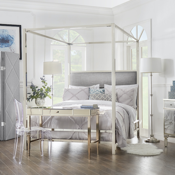 Like the previous modern canopy bed, this Metal Canopy Bed features grey linen upholstery. However, the metal frame comes in a chrome finish. The bedding comes in various shades of grey, white, and blue, for a light, clean aesthetic that complements the linen and metal of the bed. At the foot of a bed is a mirrored writing desk with an acrylic chair. The two lamps have white drum shades and metal bases, which beautifully accents the bed in the center of the room. 