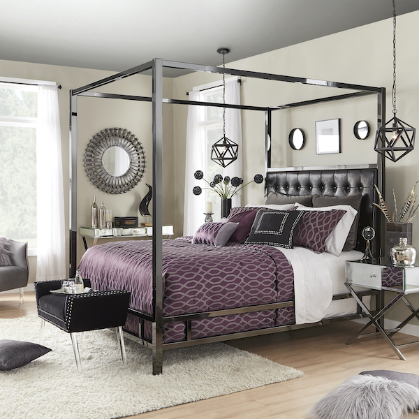 This modern canopy bed features a bonded leather upholstered headboard with plush button tufting and a black nickel finish metal frame. The bedding includes deep purple comforter, white sheets, deep purple and black pillows. This creates a rich, edgy look that is sure to make a bold statement. At the foot of the bed is a plush, black velvet bench. The nightstand is mirrored on a metal base, while a matching mirrored writing desk sits in the background.