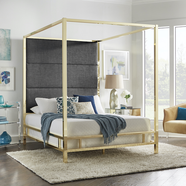 This modern canopy bed makes a bold statement in any room. The headboard is upholstered in dark grey linen while the shiny metal frame is finished in gold for a striking contrast. The bedding is primarily white, with some blue and gold blankets and pillows to complement the linen and metal. There is also a mirrored nightstand and a gold velvet accent chair. 