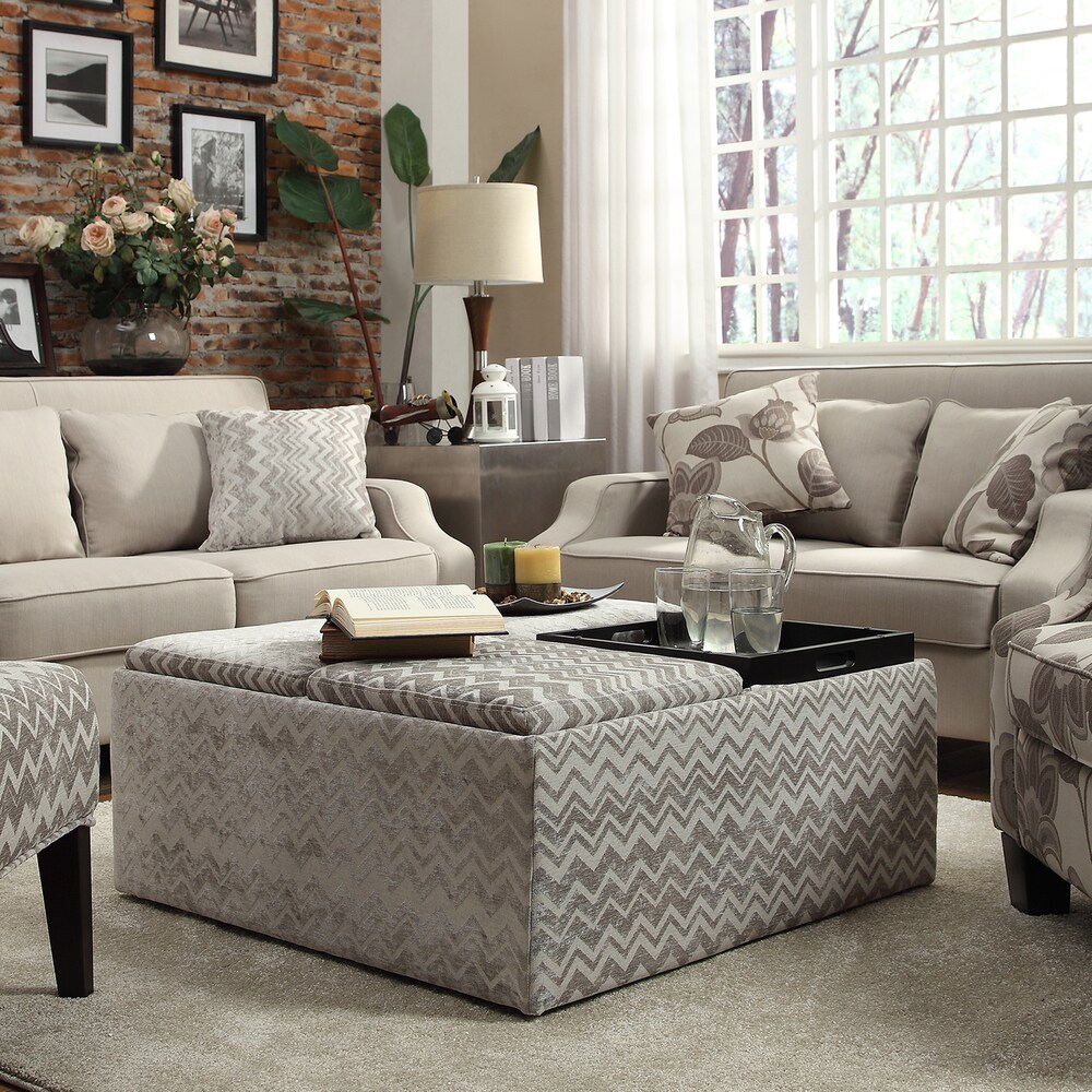 Pictured here is the Grey Chevron Storage Cocktail Ottoman by iNSPIRE Q Bold. One of the four lids on the ottoman is flipped, revealing a convenient tray to hold some glasses of water. The ottoman is also decorated with books and candle décor.