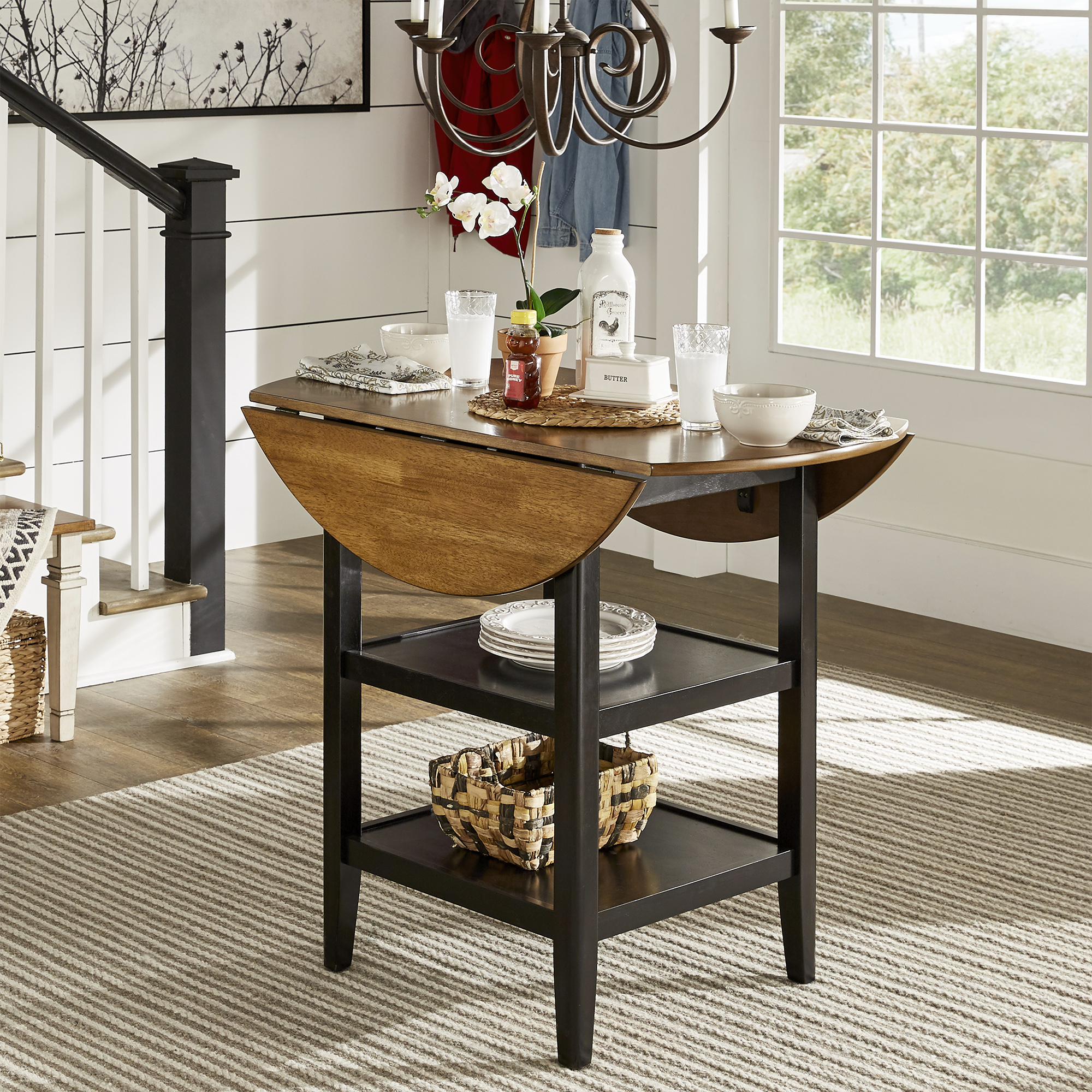 Now we move onto discussing furniture materials for hard surfaces. We begin with wood, specifically rubberwood. Pictured here is the Antique Black Drop Leaf Round Counter Height Dining Table. It has an oak-finished top with the sides dropped down for a narrower surface. The antique black finish base has two open shelves to store extra kitchen wares.