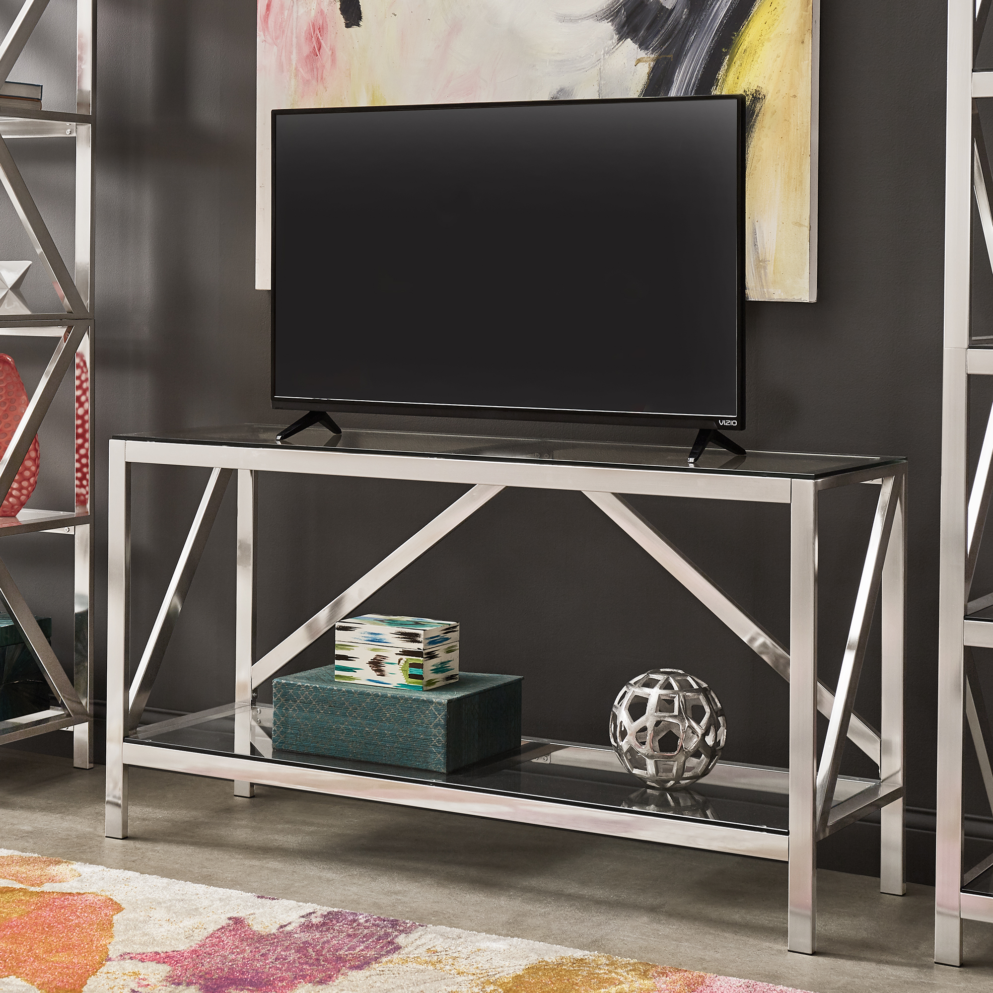This is the Brushed Nickel TV Stand by iNSPIRE Q Bold. It is depicted as a TV stand, with a TV perched on top while the lower glass shelf displays a few pieces of décor.