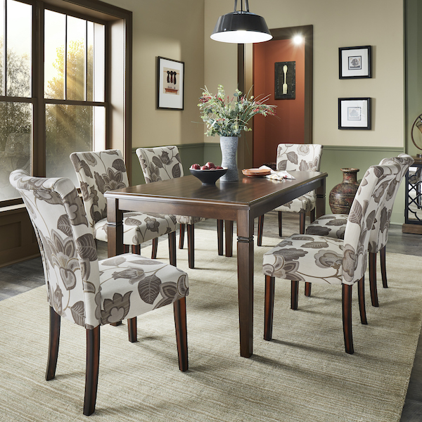 This picture is just one piece of the fall home décor trends, focusing exclusively on the dining area. There is a rich, dark wood rectangular dining table decorated with a vase of greenery and a bowl of fruit. Six parsons chairs are around the dining table, all with leaf-printed upholstery.